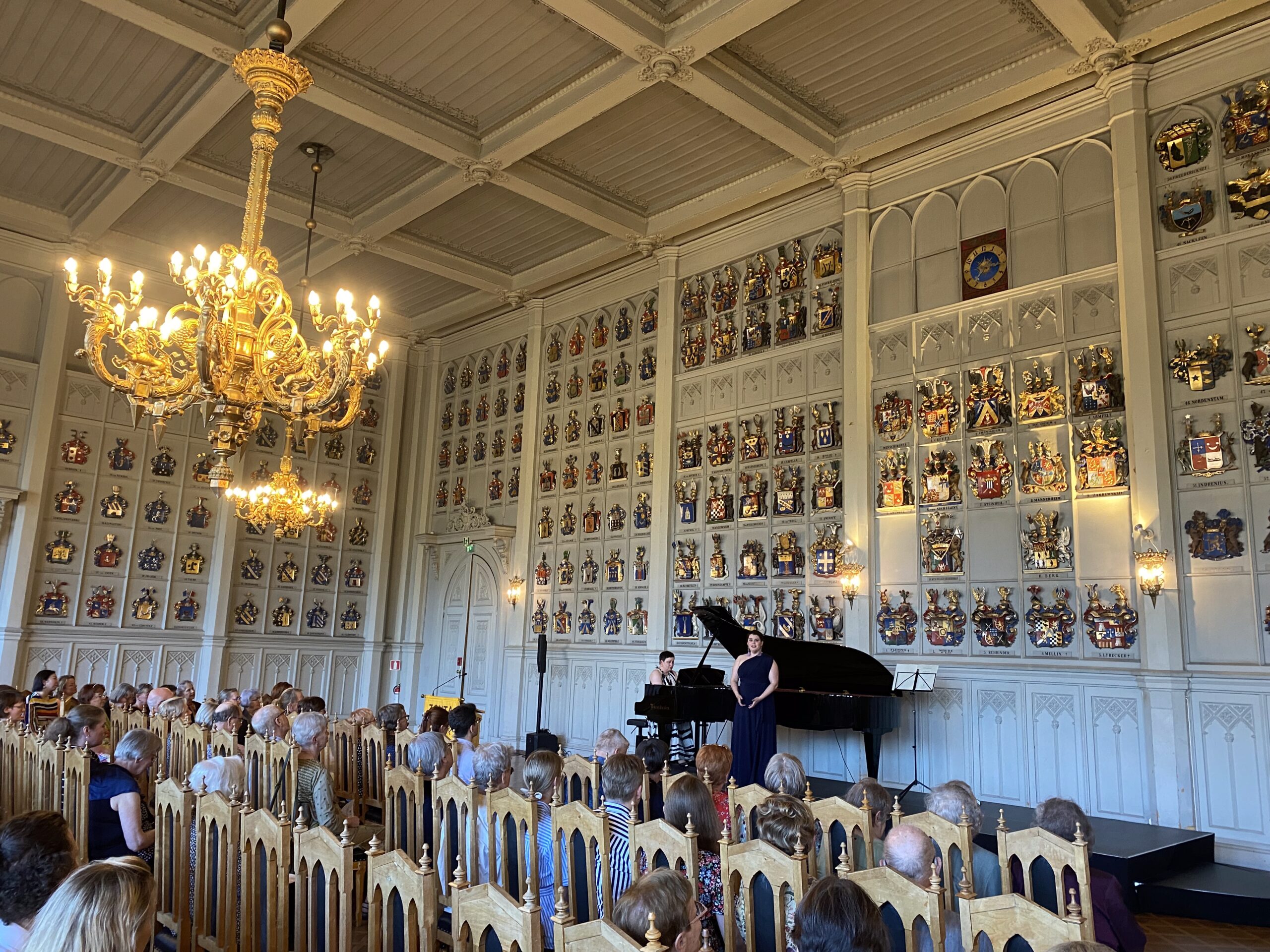 Sopraano ja pianisti esiintyvät Ritarihuoneen historiallisessa juhlasalissa aatelisvaakunoiden ympäröimänä